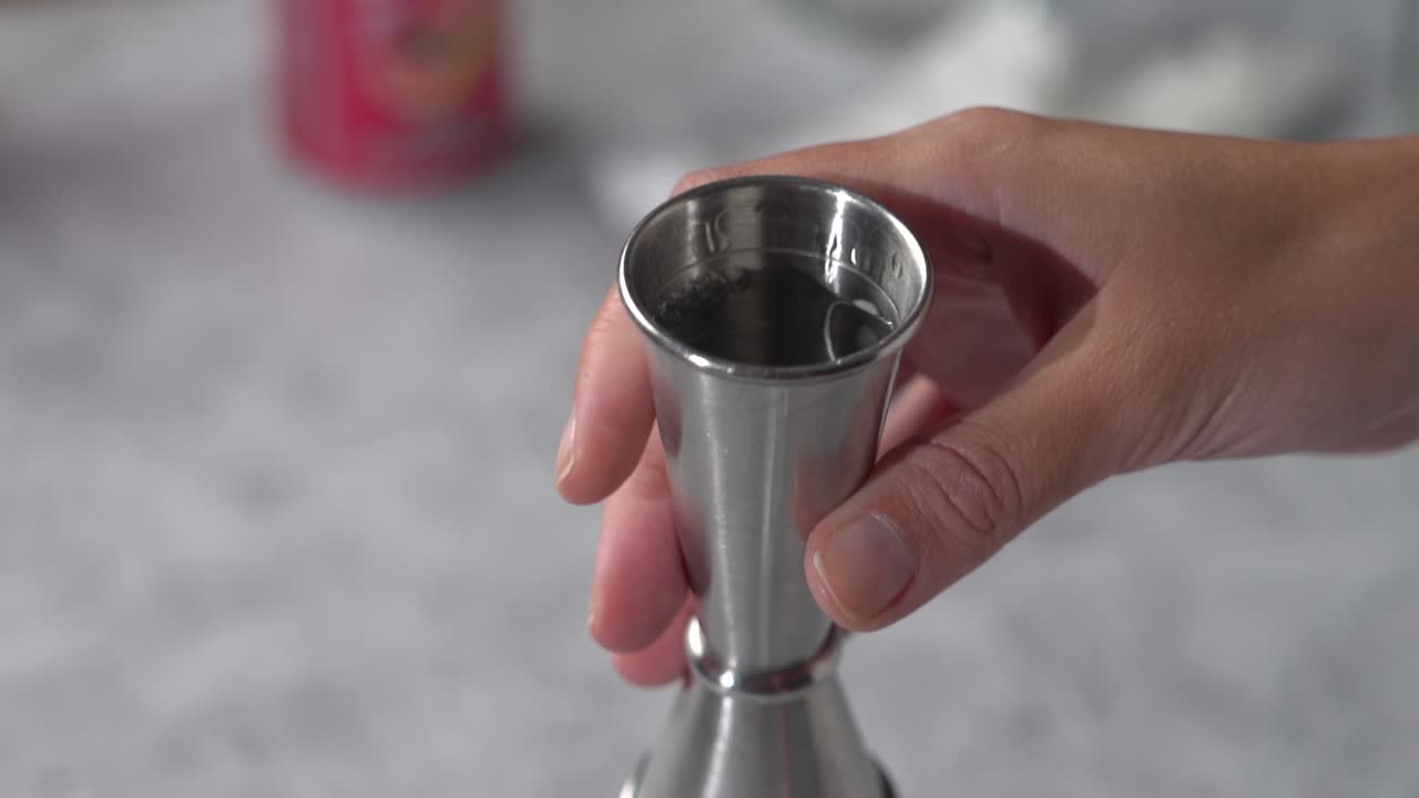 Woman pouring clear liquid from metal jigger into clear tall glass close up slow mo