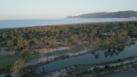 Slow-aerial-ascending-shot-along-coastline-of-Sado-River-over-forest-trees-with-view-of-sesimbra-mountains-in-backdrop