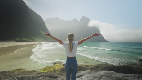 Woman-performs-yoga-sun-salutation-on-Kvalvika-Beach-in-Lofoten,-Norway,-with-mountains-in-the-background