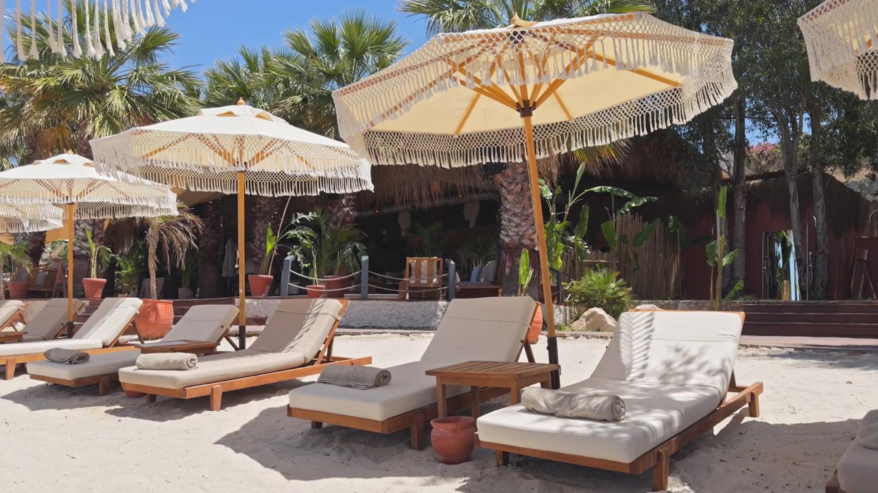 Inquadratura statica grandangolare di lettini di lusso e ombrelloni boho chic con frange allineati sulla sabbia bianca in un esclusivo resort sulla spiaggia tropicale sotto un cielo azzurro e limpido.