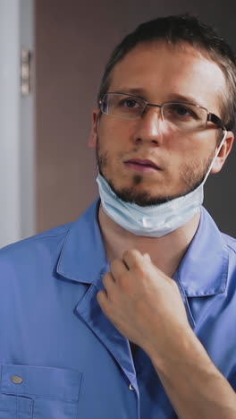 slow motion: tired man doctor in blue uniform takes off white surgical mask and glasses preparing to work near large mirror in public bathroom closeup