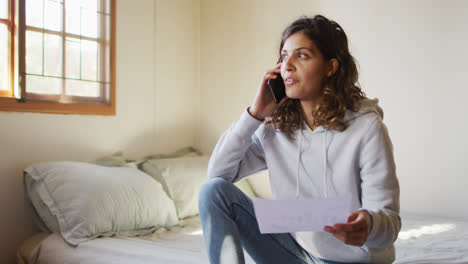 Frau-Mit-Gemischter-Rasse,-Die-Zu-Hause-Arbeitet,-Auf-Dem-Bett-Sitzt-Und-Auf-Dem-Smartphone-Spricht-Und-Papierkram-In-Der-Hütte-Hält