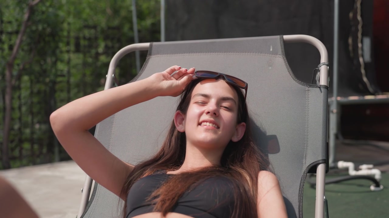 Young woman relaxes on gray outdoor lounge chair adjusting her sunglasses and lying back with long brown hair. pair of black glasses rests on wooden table beside her in outdoor setting