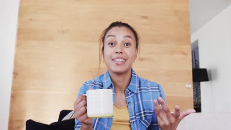 happy biracial woman making video call having coffee and talking in living room, in slow motion