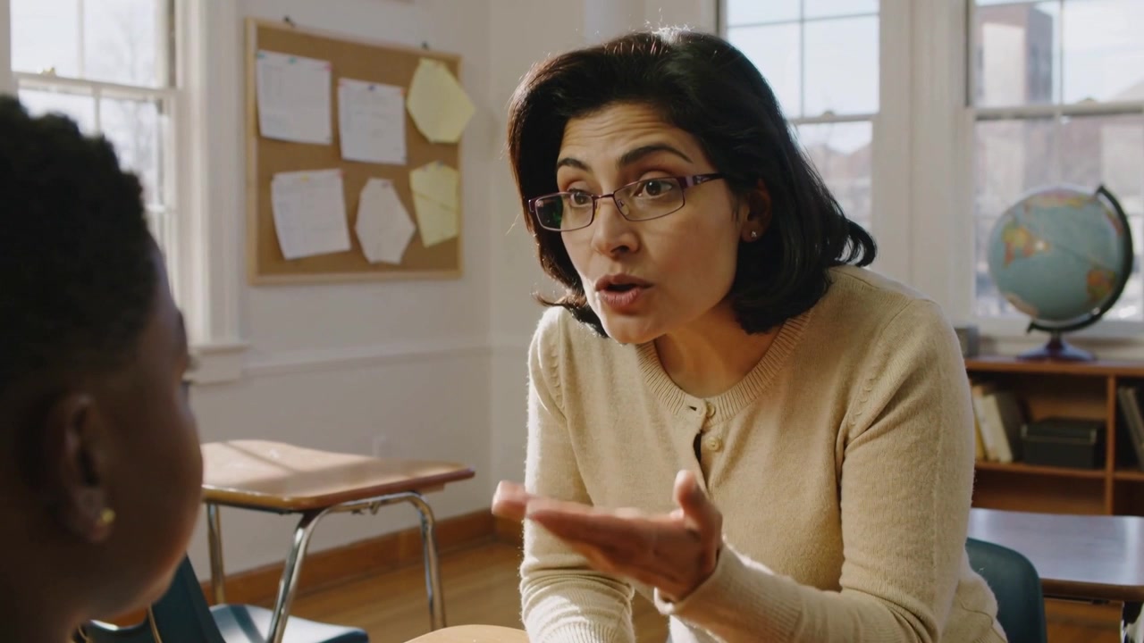 Leaning teacher with glasses beige cardigan starting talking for help, gesturing at desk with globe