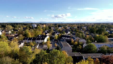 Vista-Aérea-De-Los-Suburbios-De-Munich:-Drones-Se-Deslizan-Mostrando-Nítidos-Bosques-Otoñales-Que-Conducen-A-Majestuosos-Fondos-Montañosos,-Una-Armonía-Urbano-natural