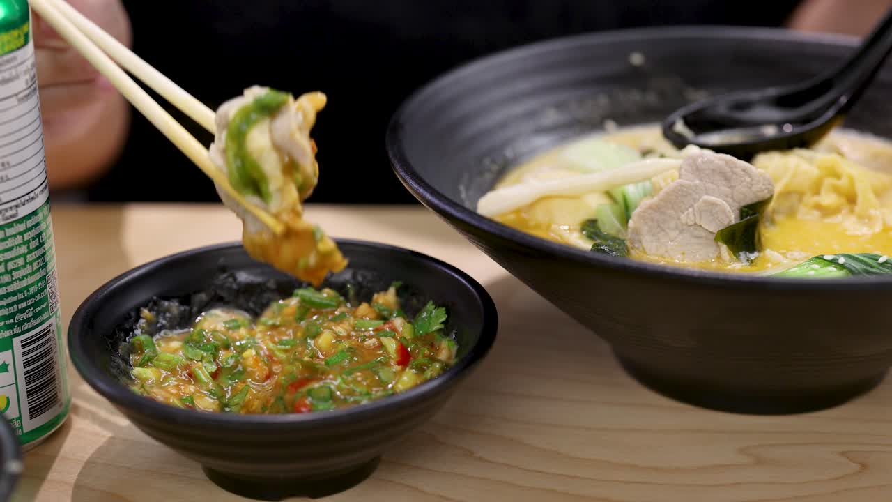 Hand uses chopsticks to dip noodle soup toppings in spicy sauce, bright tabletop lighting, closeup
