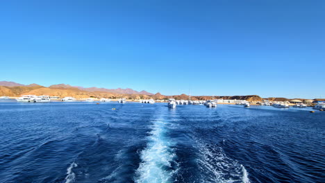 Hermosos-Cielos-Azules-Desde-La-Vista-Trasera-De-Un-Barco-Que-Viaja,-Dejando-Atrás-El-Puerto-Con-Barcos-De-Lujo-Al-Fondo.