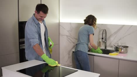 Young-smiling-married-couple-in-casual-clothing-and-both-in-green-gloves-cleaning-kitchen-after-moving-to-new-home,-household.-Woman-is-washing-the-dishes,-man-is-cleaning-a-kitchen-stove-with-a-mop-and-chemical-spray