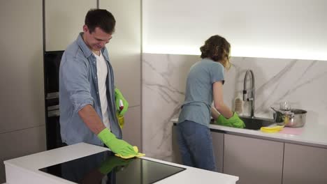 young smiling married couple in casual clothing and both in green gloves cleaning kitchen after moving to new home, household. woman is washing the dishes, man is cleaning a kitchen stove with a mop and chemical spray