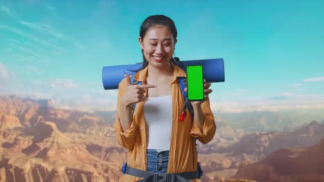 asian female hiker with mountaineering backpack smiling and pointing to mock up green screen smartphone while traveling at the top of mountain
