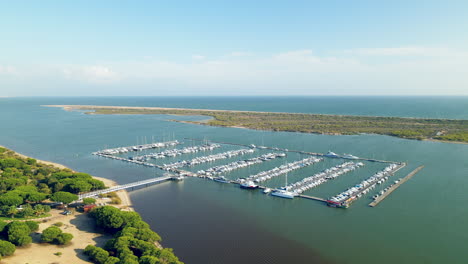 Retiro-Aéreo-De-Los-Muelles-De-Puerto-Marina-El-Rompido-Con-Muchos-Yates,-Veleros,-Embarcaciones-Y-Muelle-En-El-Río-Piedras,-España,-Océano-Atlántico-En-El-Fondo-Detrás-Del-Asador