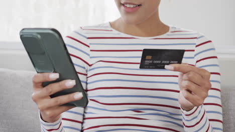 happy biracial woman sitting on sofa using smartphone for online shopping, slow motion