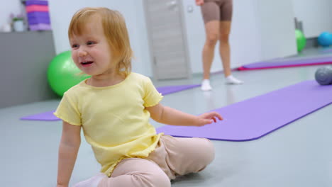 smiling little girl in yellow shirt playing with blue ball on gym floor, reaching for it as it rolls away, with woman in brown outfit standing in background near workout mats and fitness equipment