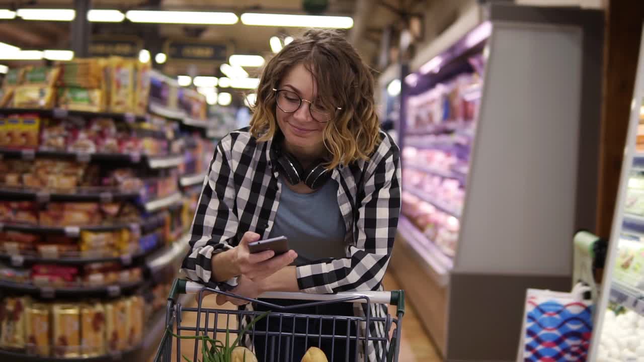 compras, tecnología, ventas, consumismo y concepto de personas - mujer con teléfono inteligente y auriculares en el cuello caminando en el supermercado. mujer sonriente enviando mensajes de texto con amigos, apoyada en su carrito de compras