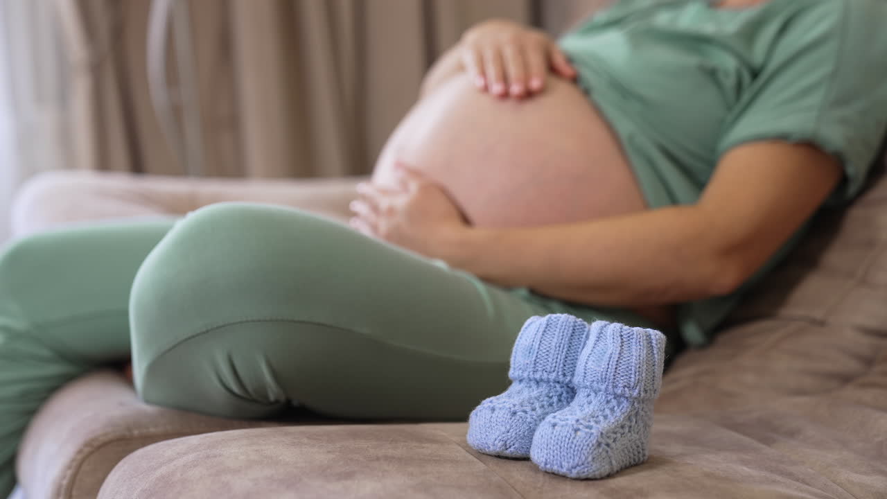 Pregnant woman with big bare belly sitting on the couch. Lady caressing her pregnant belly tenderly and then putting baby socks on it.