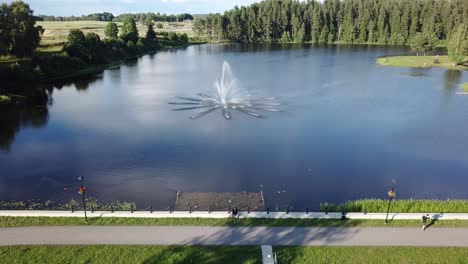 Hombre-Sentado-Al-Lado-De-Teperis,-Hermoso-Lago-Con-Fuente-En-Un-Antiguo-Parque-En-Smiltene---Dron-Ascendente,-Retroceso