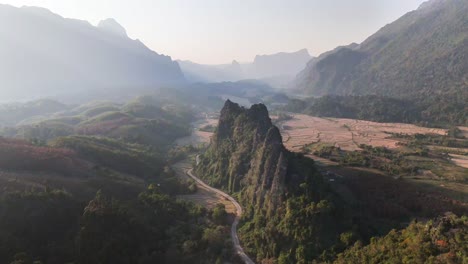 dramatic limestone formations rise from the valley floor in nam xay, vang vieng, laos, surrounded by rolling hills, farmland, and a winding rural road in soft morning or evening light