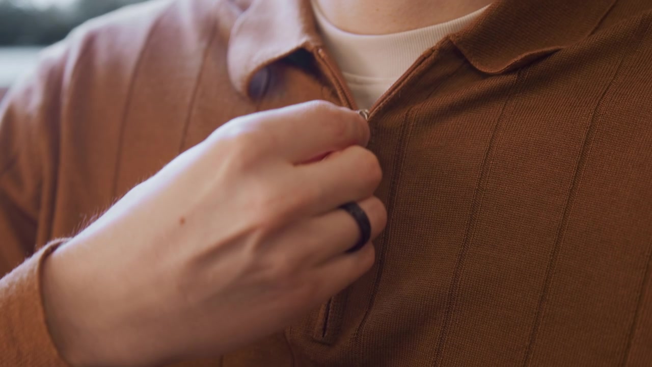 Close up of man zipping collar on orange ribbed shirt with soft lighting and natural background. hand wearing black ring pulls down to fasten zipper while details like fabric texture