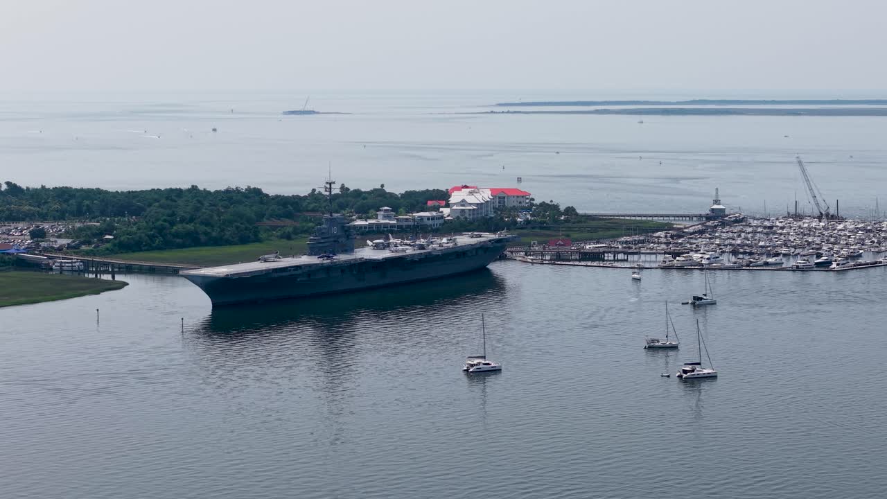 A high-altitude drone captures a wide shot of the USS Yorktown aircraft carrier docked at Patriots Point, surrounded by calm harbor waters and small boats