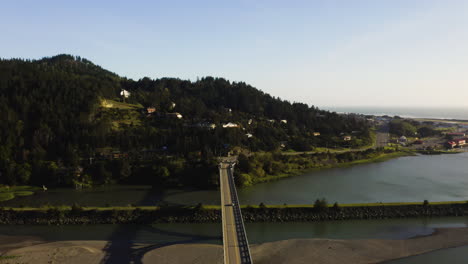 Drone-flying-backwards-over-the-Rogue-River-bridge-in-Gold-Beach,-Southern-Oregon,-revealing-cars-crossing-the-large-span