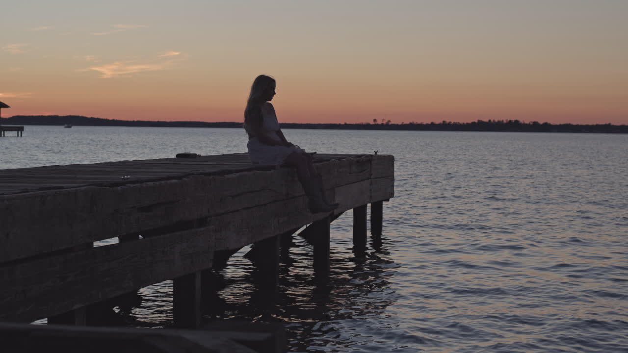 Bella ragazza seduta sul bordo di un molo al tramonto con il vento che soffia