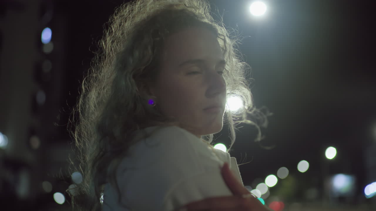 Young woman standing outside during cold night with hand on shoulder, illuminated by glowing street lights in urban environment, her face calm and thoughtful against blurred background of city bokeh