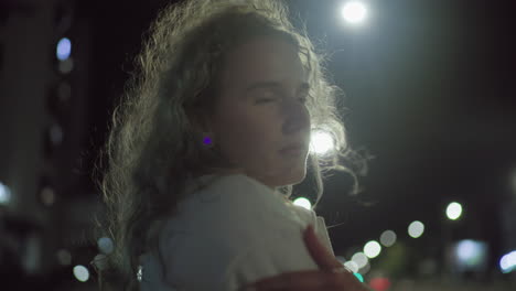 young woman standing outside during cold night with hand on shoulder, illuminated by glowing street lights in urban environment, her face calm and thoughtful against blurred background of city bokeh