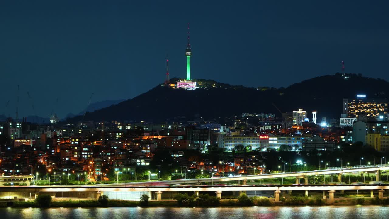 Night timelapse of Seoul showing the illuminated Namsan Tower and the bustling Yongsan district, with streaks of car headlights from fast-moving traffic along the Gangbyeonbuk-ro expressway