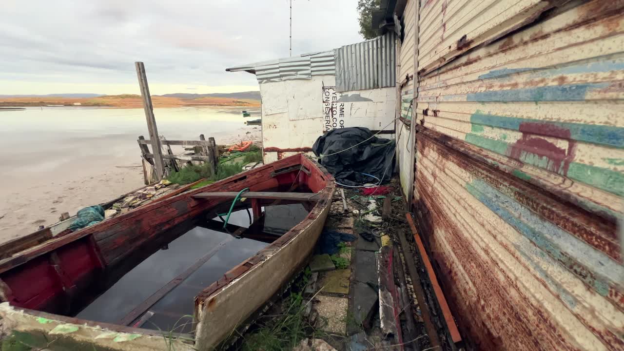 Lento retroceso a lo largo de una cabaña de pescadores oxidada que revela un barco abandonado lleno de agua cerca del estuario de Barbate, Cádiz