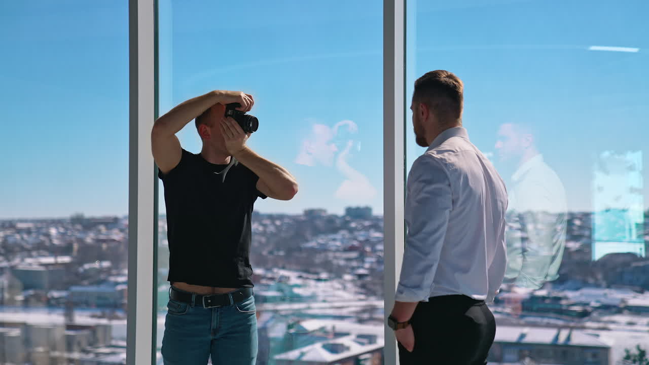 Young photographer taking photos of a businessman. Man shooting handsome man model in white shirt near windows. Working with camera. Profile view.