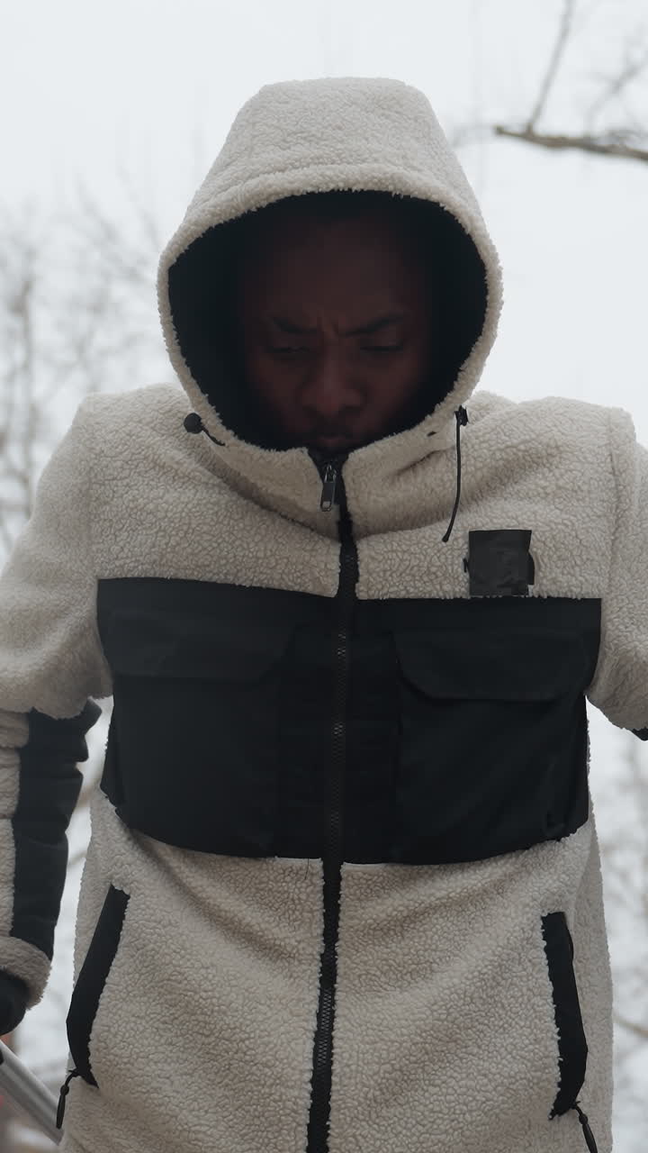 젊은이가 겨울 동안 야외 평행 바에서 근육을 쌓기 위해 푸시업 운동을 수행하는 클로즈업, 그는 후드 재과 장갑을 입고 있으며, 차가운 공기에서 눈에 띄는 숨을 쉬고 있습니다.