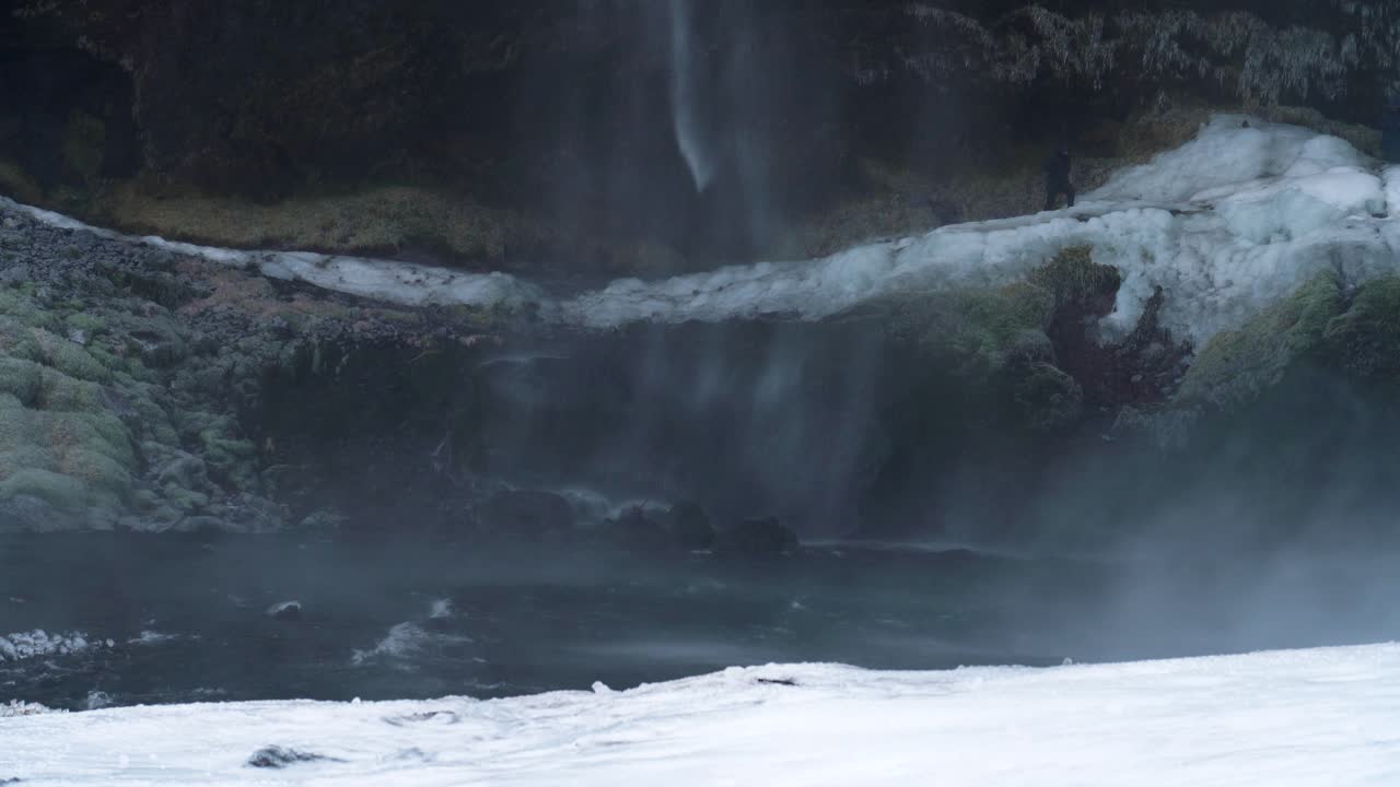 아이슬란드에서 안개가 자욱한 스프레이와 함께 폭포 스트림에 의해 얼음 풍경에 서있는 관광객