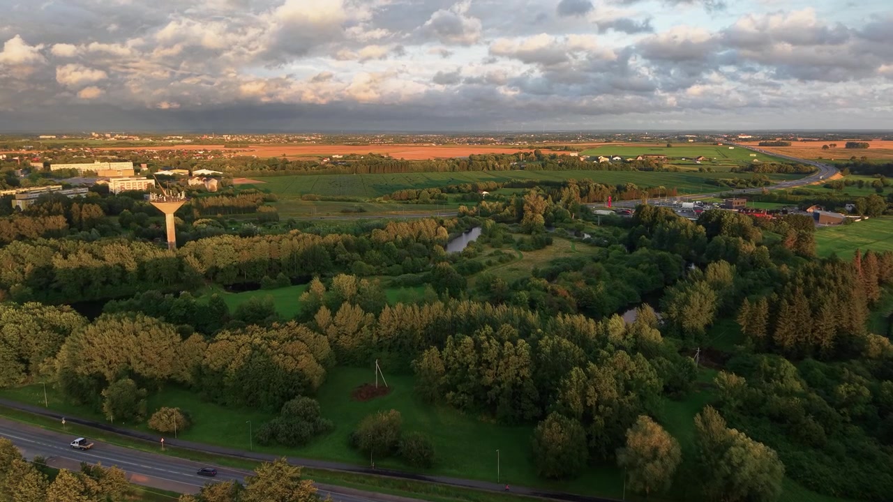 Dronevideo tijdens het gouden uur zweeft boven weelderig platteland, dichte bossen, een kronkelende rivier, verre landbouwgrond en landelijke wegen onder dramatische wolken en warm natuurlijk zonlicht, met levendige landschapsdetails