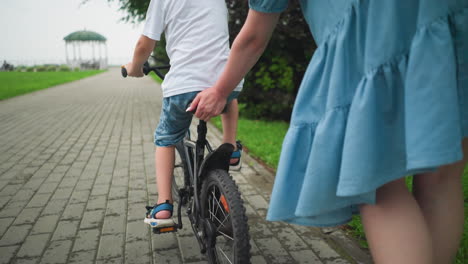 a woman holds the seat of her son s bicycle, helping him maintain balance as they ride along a paved walkway surrounded by greenery