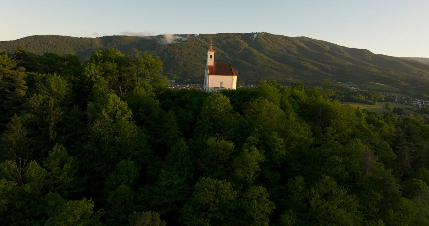 Veduta aerea di una chiesa isolata in cima a una collina sopra una fitta foresta e dolci montagne in Slovenia, durante la calda luce del tramonto e una serena atmosfera serale.