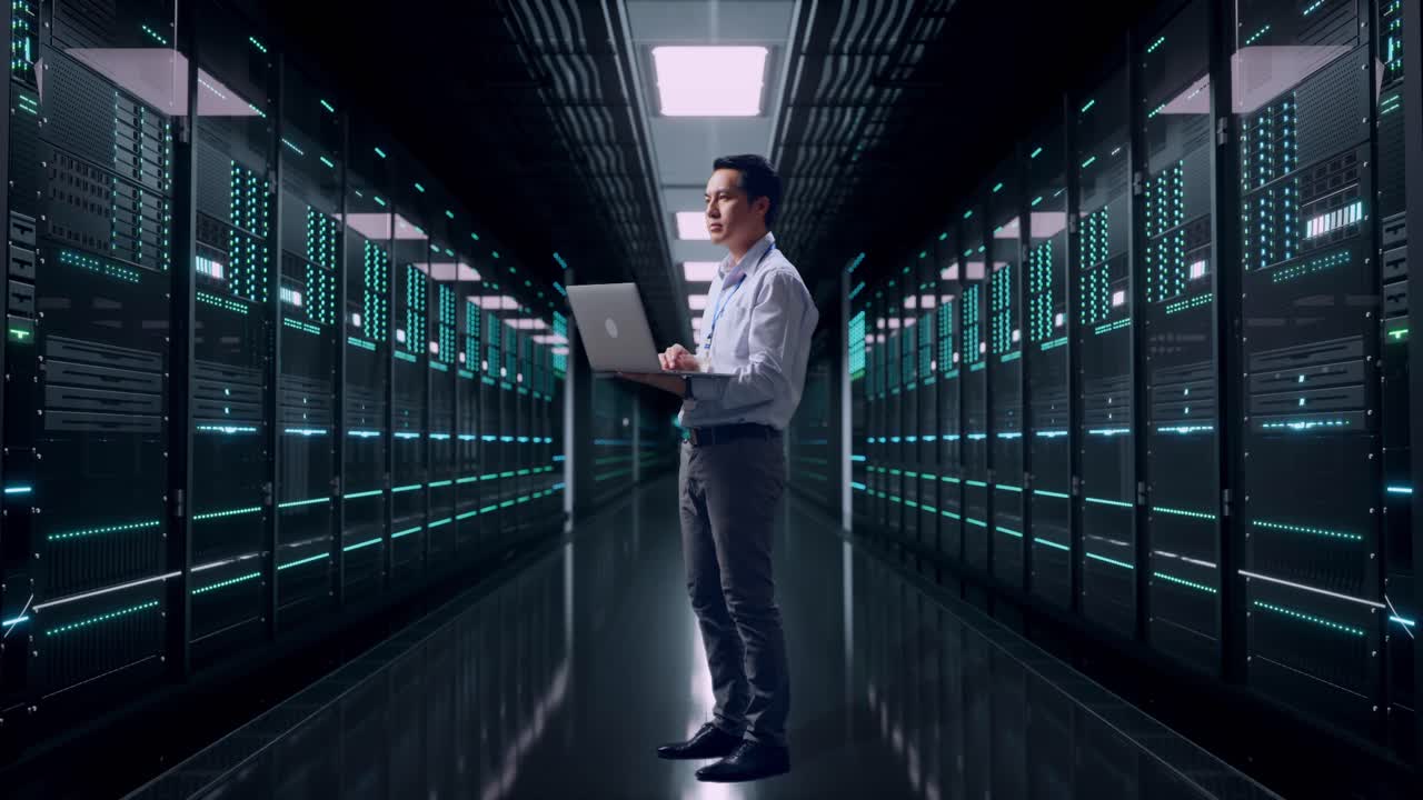 Helkropsbillede af en asiatisk professionel mandlig medarbejder set fra siden i et serverrum i et datacenter, kigger opmærksomt op, før han koncentrerer sig om den bærbare computer og fortsætter med at taste.