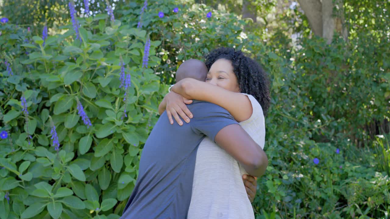 Coppia afroamericana che si guarda negli occhi, stringendosi in un tenero abbraccio in un giardino tra fioriture viola, smartwatch, magliette.