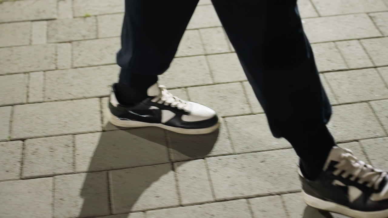 Leg view of individual in black and white sneakers walking along interlocked concrete pavement with evening shadows cast on ground and fresh green grass sprouting between tiles