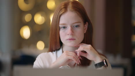 thoughtful brooding remote working red haired woman sitting infront of a laptop or notebook in casual outfit on her work desk in her modern airy bright living room home office with many windows