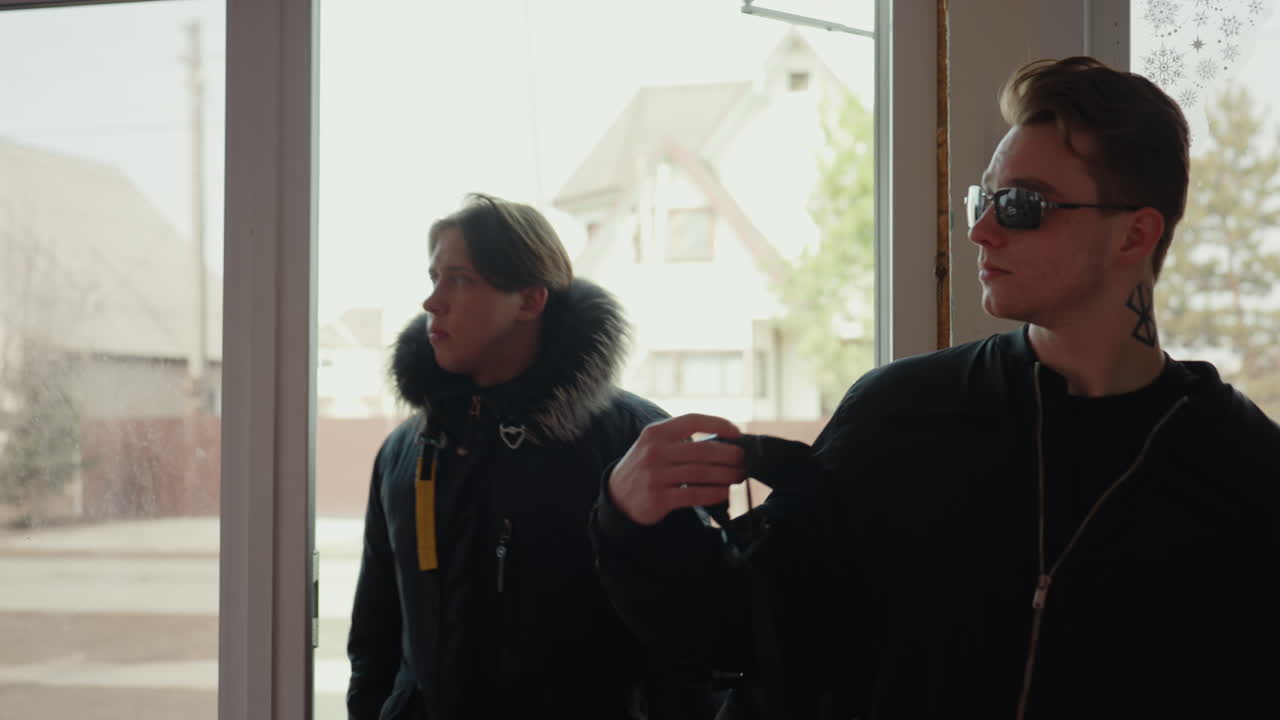 Two guys dressed in dark jackets enter through open glass door, one wearing sunglasses and backpack, other in fur hooded coat, bright daylight and parked car visible outside suburban street background