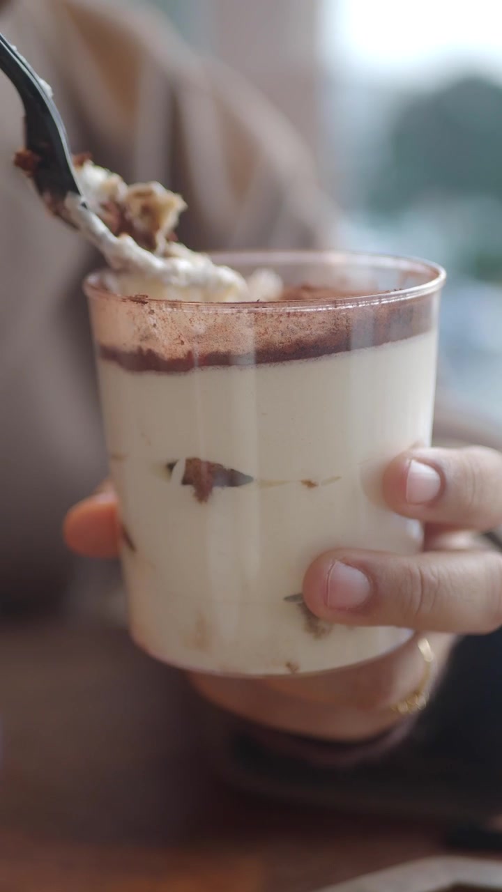 Primo piano di una mano che mangia tiramisù da una coppetta di plastica.