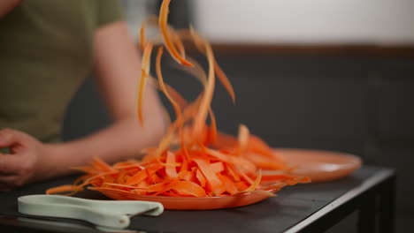 close up of hand lifting and dropping peeled carrot strips back onto orange plate, white peeler resting on black table surface, partial figure in green shirt visible in soft kitchen background