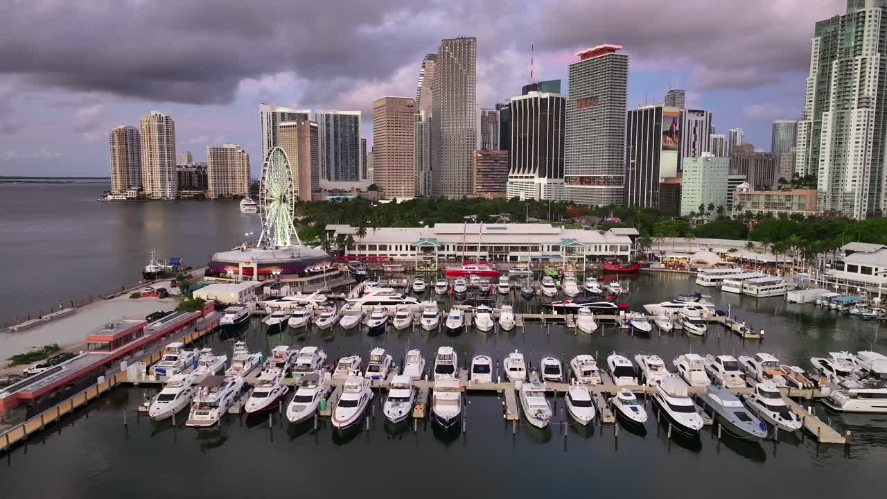 Yachts docked at the marina near Bayside Marketplace in Downtown Miami with the city skyline and Ferris wheel in the background. The view looks across Biscayne Bay. Aerial.