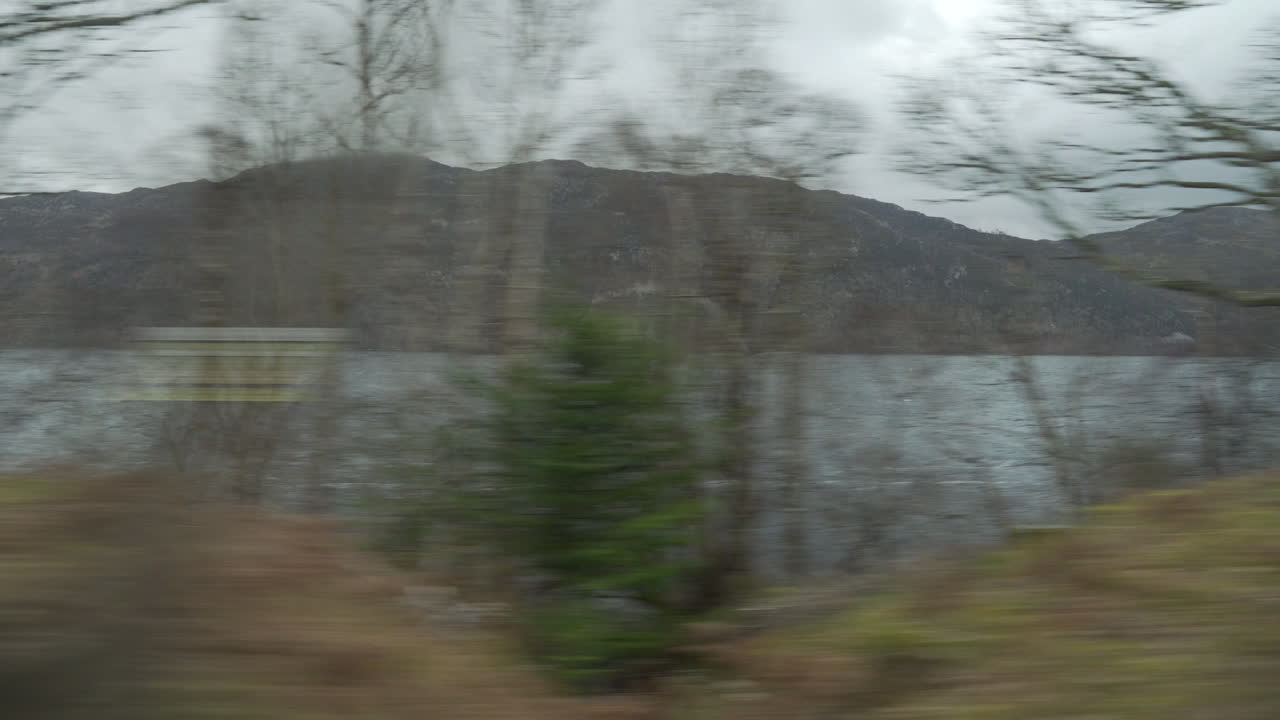 Steady moving shot from vehicle with trees passing by with wonderful dramatic sunset light and nice clouds with Loch Ness lake in the background in Scotland