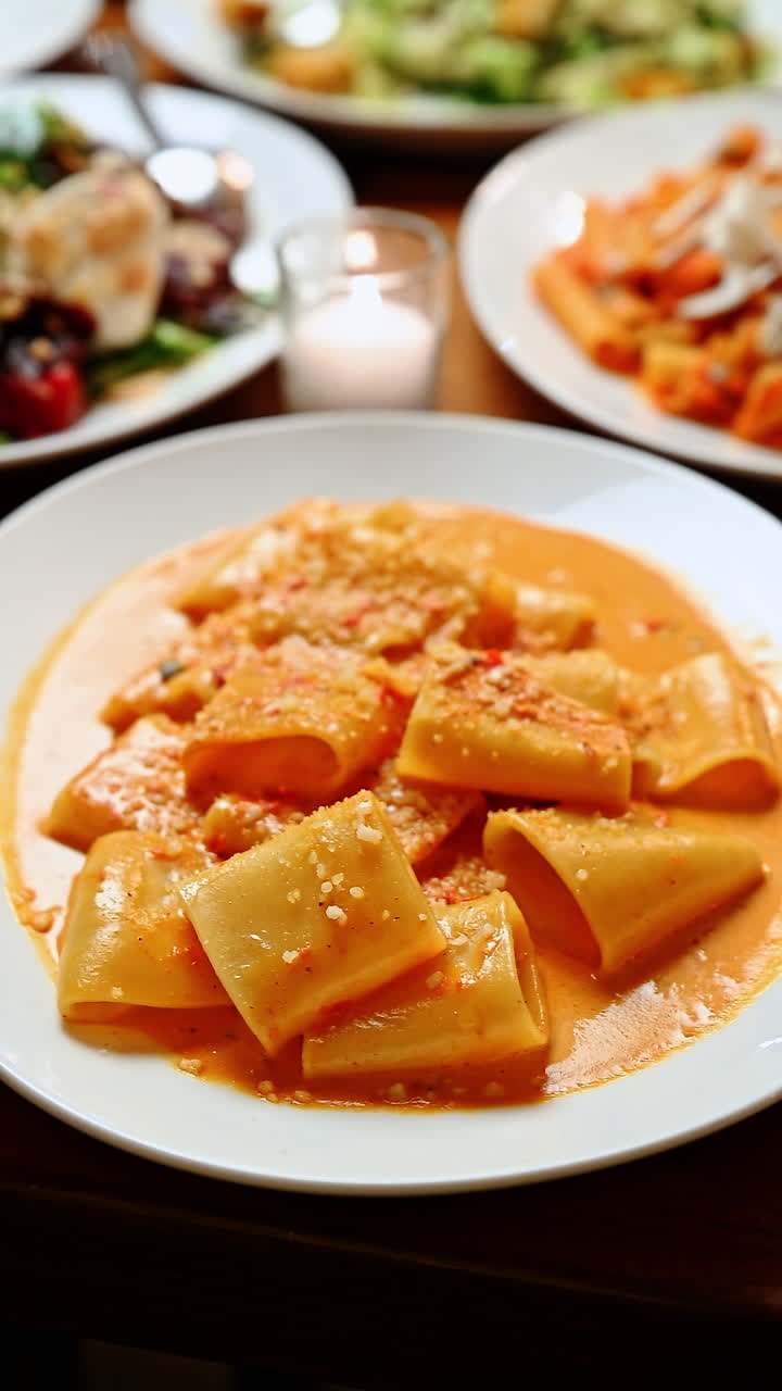 Pasta servita in un piatto bianco con salsa e formaggio in un ristorante. Un piatto di pasta con salsa arancione e formaggio è mostrato su un tavolo con contorni nelle vicinanze.