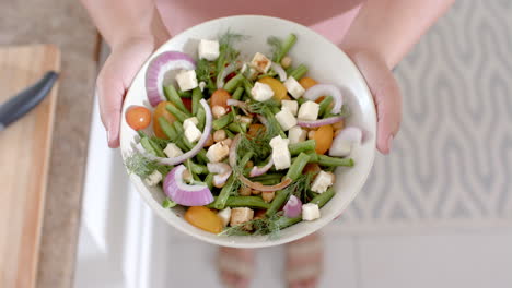 overhead hands of biracial woman holding bowl of feta salad in kitchen, copy space, slow motion