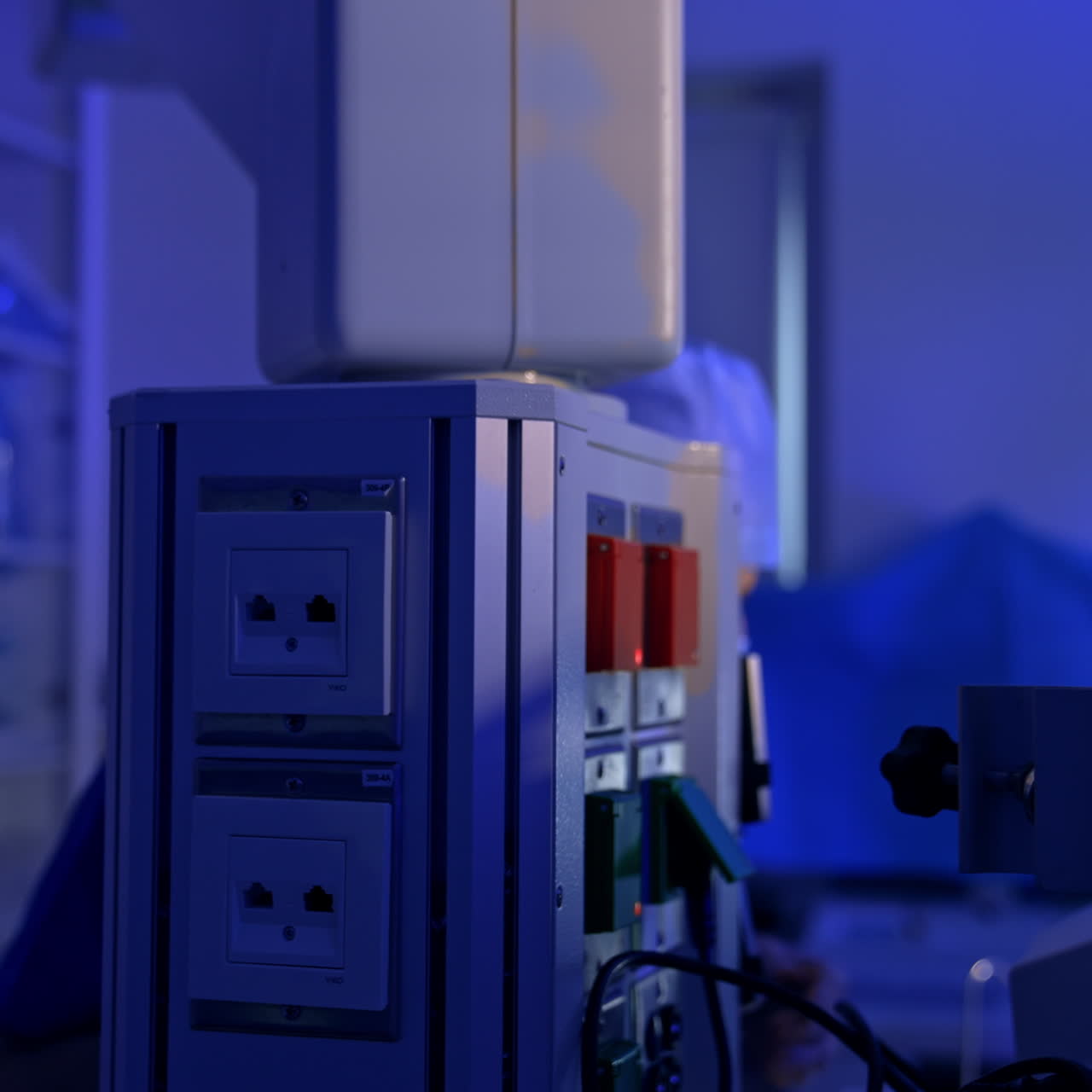 Male medic in mask and cap plugs the equipment in modern surgery room. Operational room is illuminated blue