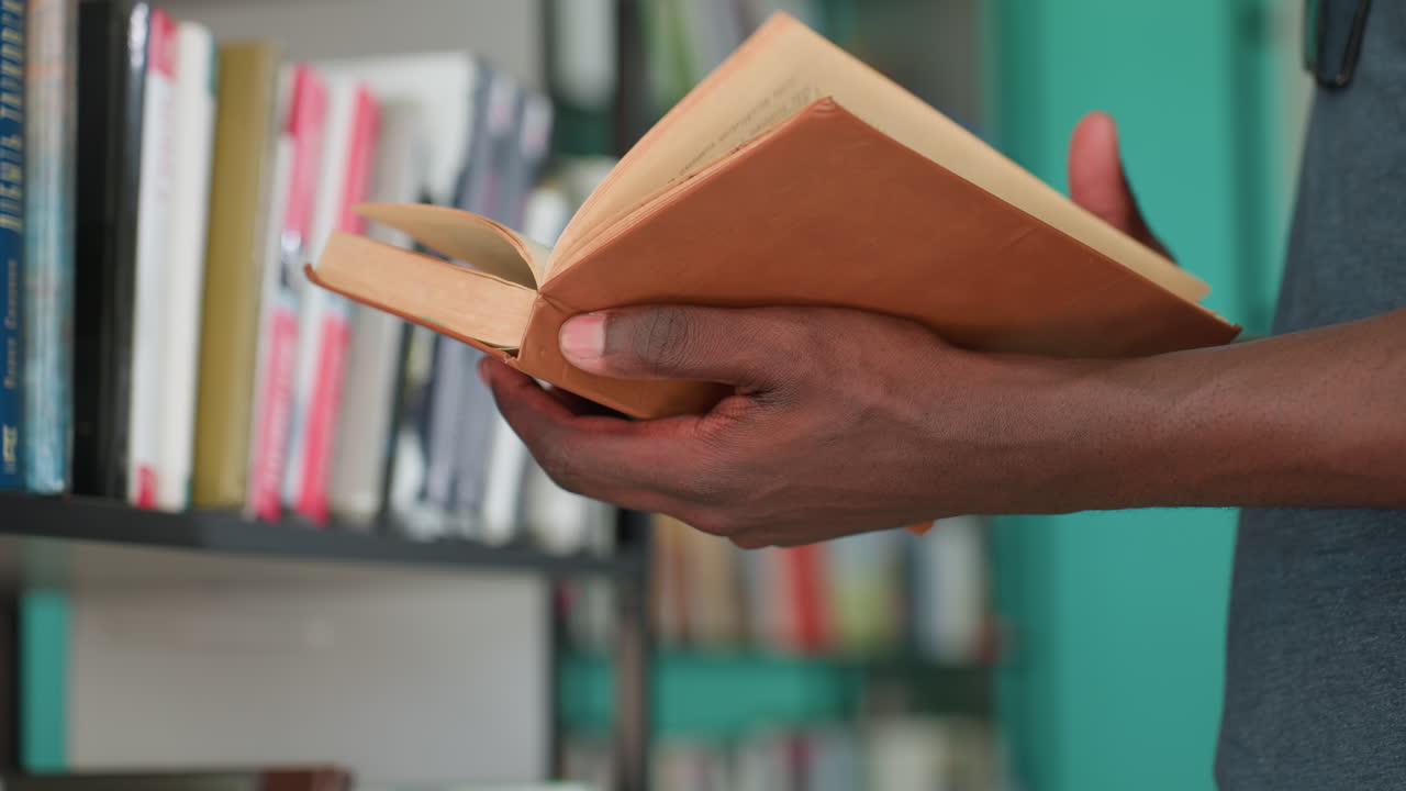 Close up of person holding and flipping through old book in library, focusing on hands and worn pages with faded ink, among blurred shelves in background