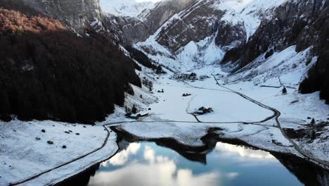 Aerial-flyover-above-Seealpsee-in-Appenzell,-Switzerland-in-winter-with-pan-up-from-the-a-reflection-on-the-lake-towards-the-mountains-of-Alpstein